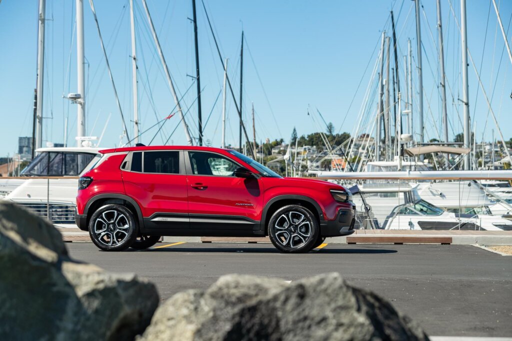 Side profile of the Jeep Avenger, pitcured in red, parked behind rocks at an Auckland marina