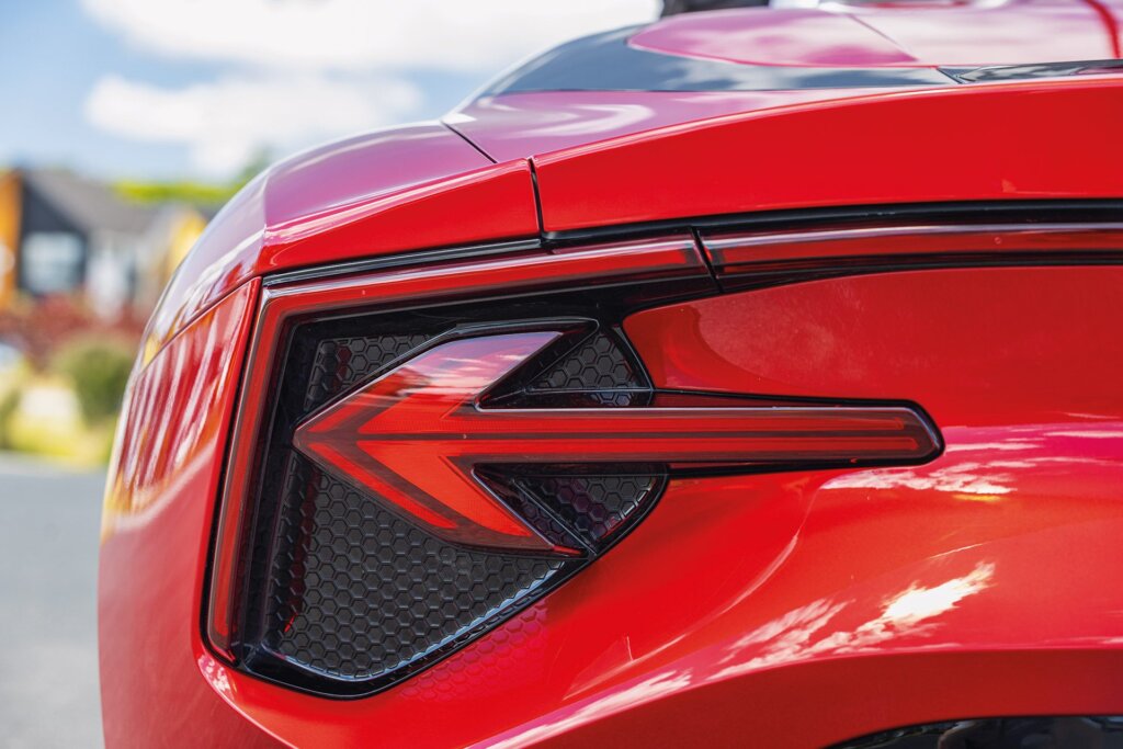 Close-up of the rear left taillight of a red sports car with a distinctive arrow-shaped design and red and black accents, reflecting the surroundings