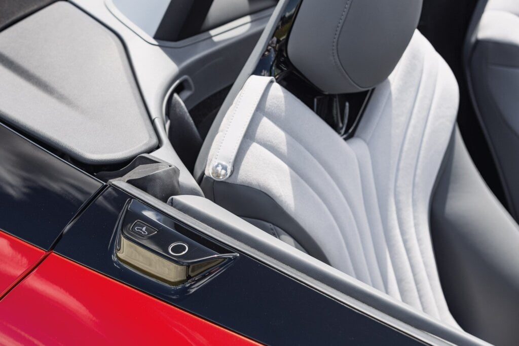 Convertible car interior showcasing a light grey fabric passenger seat with a modern, contoured design. The control panel on the side door features two buttons for seat adjustments. The car door has a glossy black and red finish