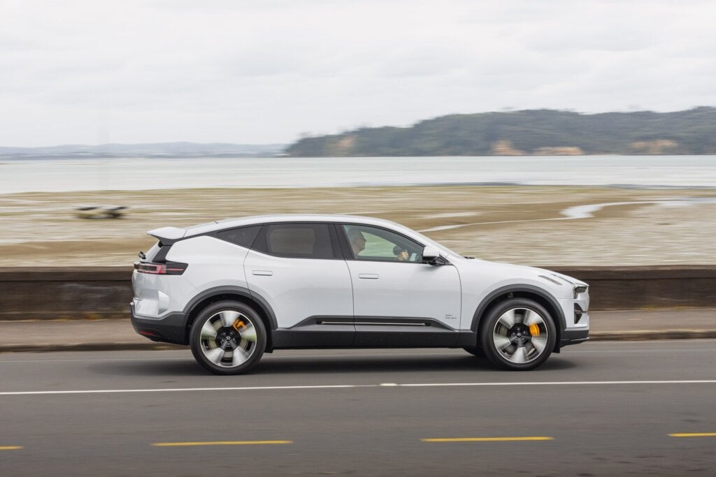 A white Polestar 3 electric SUV driving along a coastal road, showcasing its sleek design and scenic waterfront background